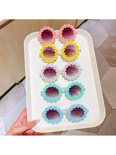 A white tray of sunglasse being held by a woman's left hand. The sunglasses pictured have daises surrounding the lenses in a circle. Each pair is a different color of flowers: pink, yellow, white, mint, and blue. The flowers have a yellow or orange center. 